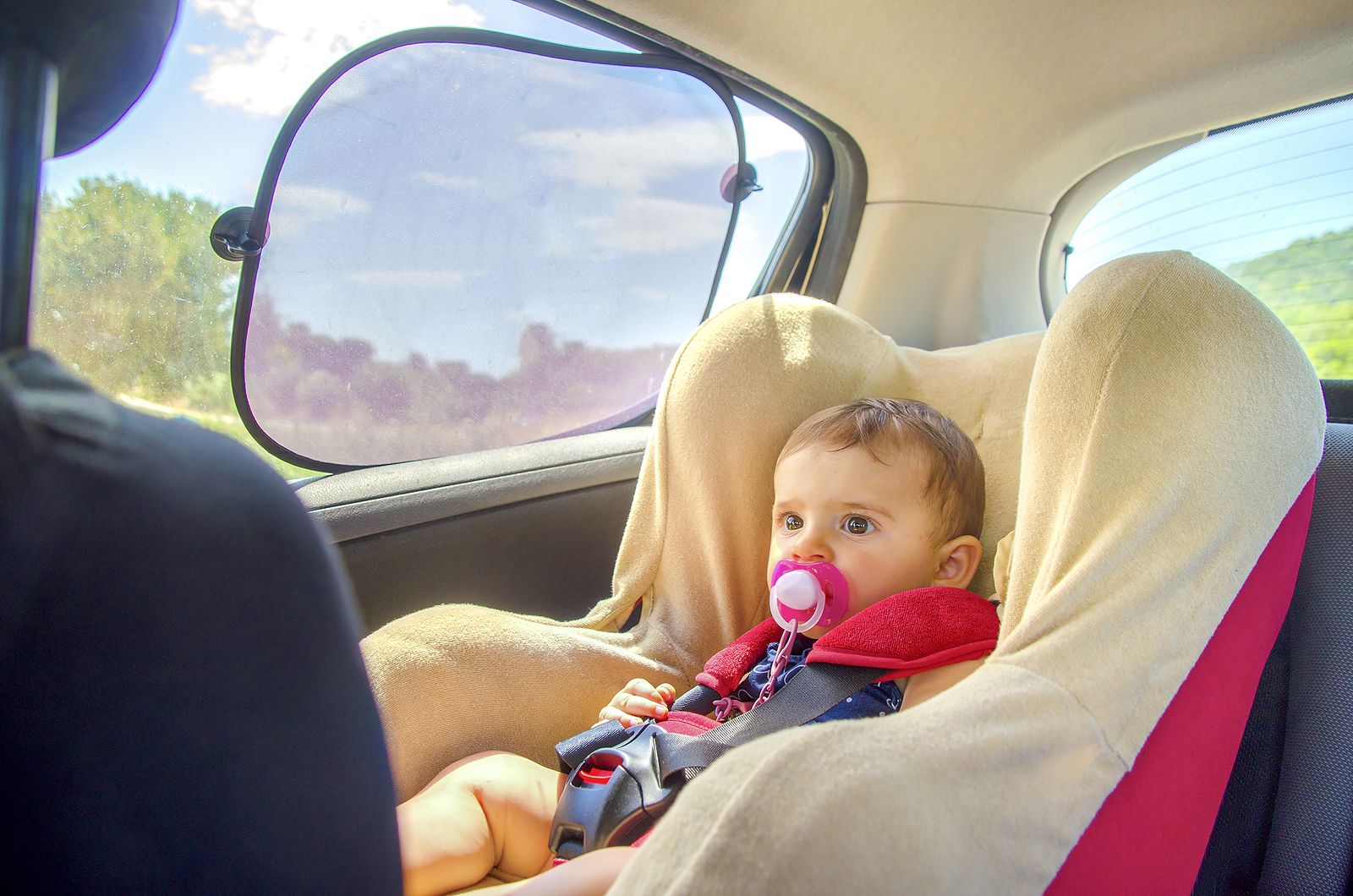 Baby with pink dummy in car sear next to window with sunshade